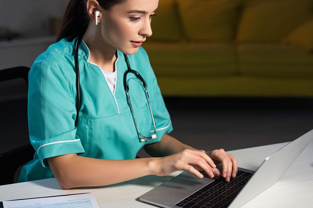 enfermeira atraente em uniforme sentado à mesa e usando laptop durante o turno da noite
 - Foto, Imagem