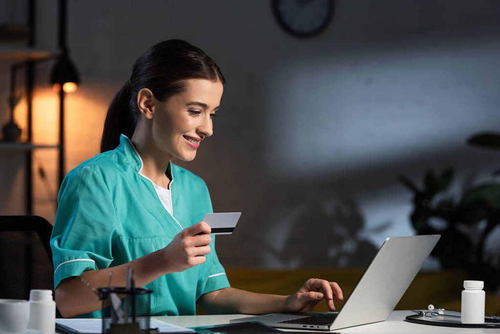 glimlachende verpleegster in uniform verpleegster in uniform met creditcard en laptop tijdens de nachtdienst - Foto, afbeelding