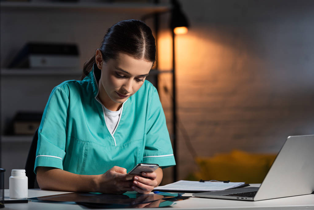enfermeira atraente em uniforme sentado à mesa e usando smartphone durante o turno da noite
 - Foto, Imagem