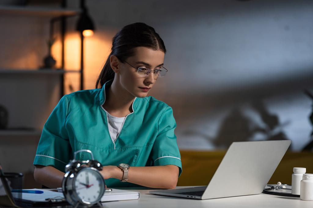 enfermera atractiva en uniforme y gafas sentadas en la mesa y mirando a la computadora portátil durante el turno de noche
 - Foto, Imagen