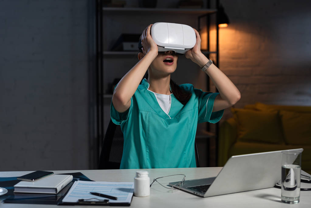 geschokt verpleegkundige in uniform met virtual reality headset zittend aan tafel tijdens nachtdienst - Foto, afbeelding