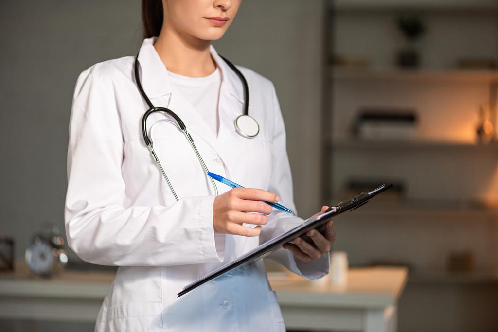 bijgesneden weergave van arts in witte jas schrijven in klembord tijdens de nachtdienst - Foto, afbeelding