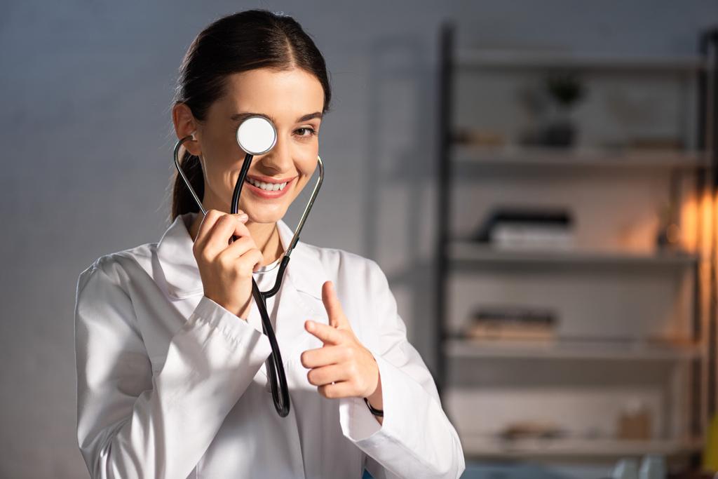 χαμογελαστός γιατρός με λευκό παλτό κρατώντας στηθοσκόπιο και δείχνοντας με το δάχτυλο κατά τη διάρκεια της νυχτερινής βάρδιας - Φωτογραφία, εικόνα