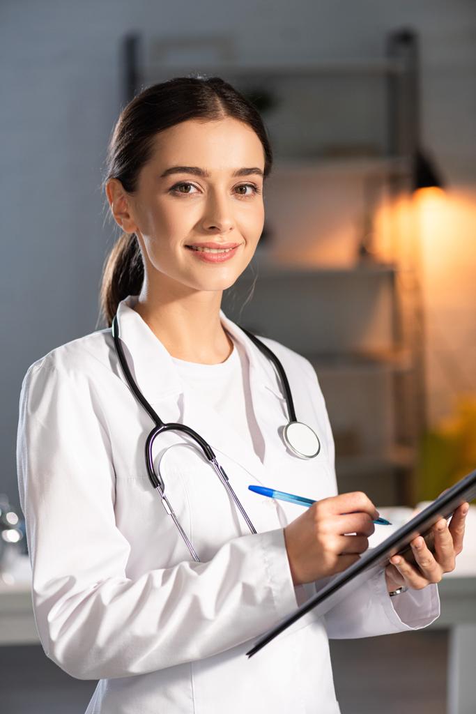 attraktiver und lächelnder Arzt in weißem Mantel mit Stethoskop, das Stift und Klemmbrett während der Nachtschicht hält - Foto, Bild