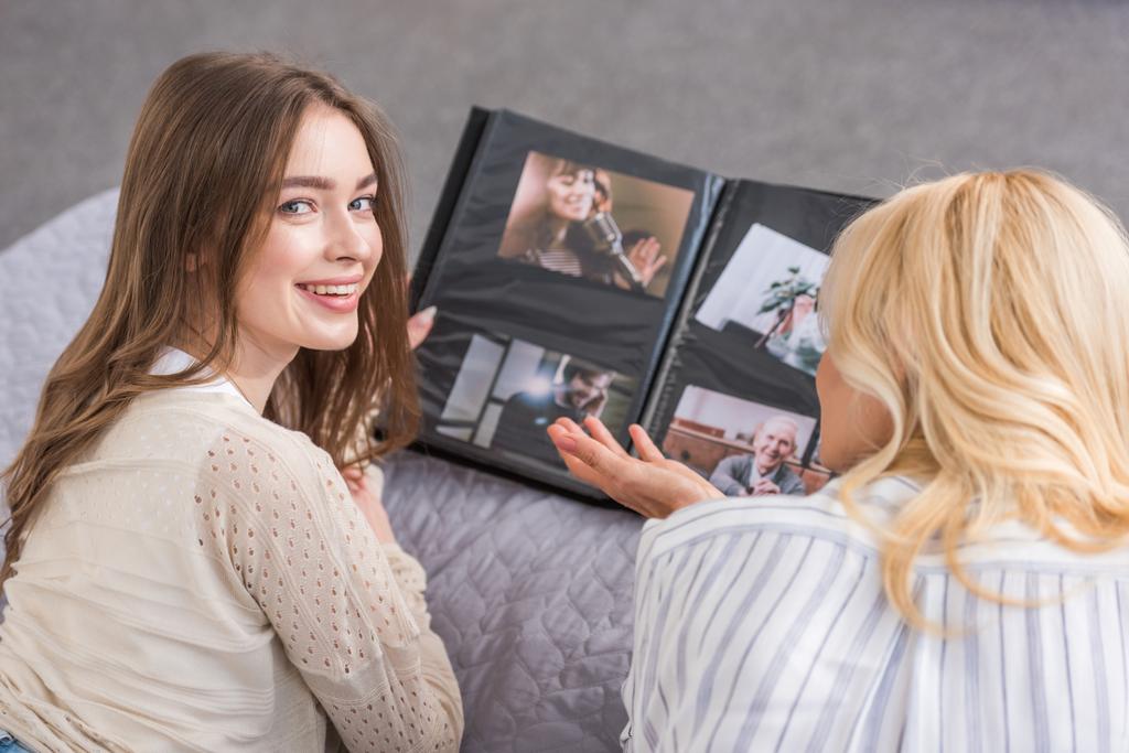 cheerful girl looking at camera while lying on bed near mother looking at photo album - Photo, Image