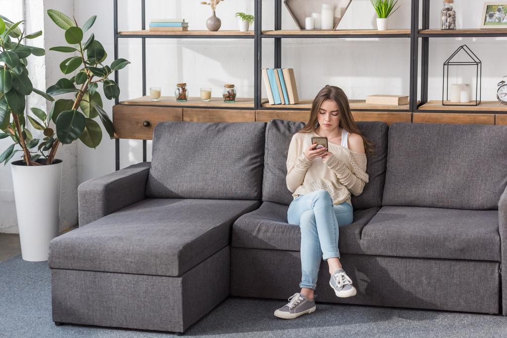 όμορφη κοπέλα χρησιμοποιώντας smartphone, ενώ κάθεται στον καναπέ στο σαλόνι - Φωτογραφία, εικόνα