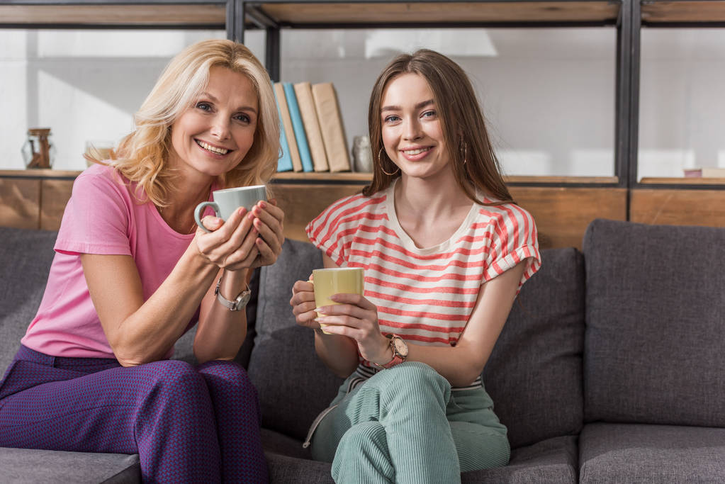 明るいお母さんと娘はソファに座ってお茶を飲みながらカメラを見て - 写真・画像