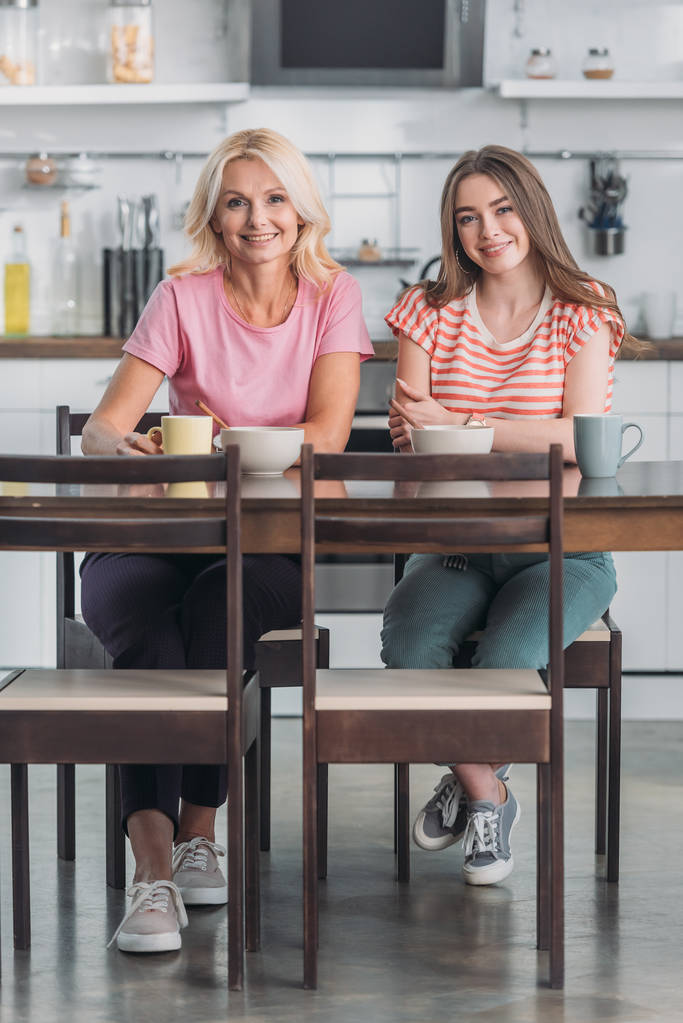 glimlachende moeder en dochter kijken naar camera terwijl ze aan de keukentafel zitten en ontbijten - Foto, afbeelding