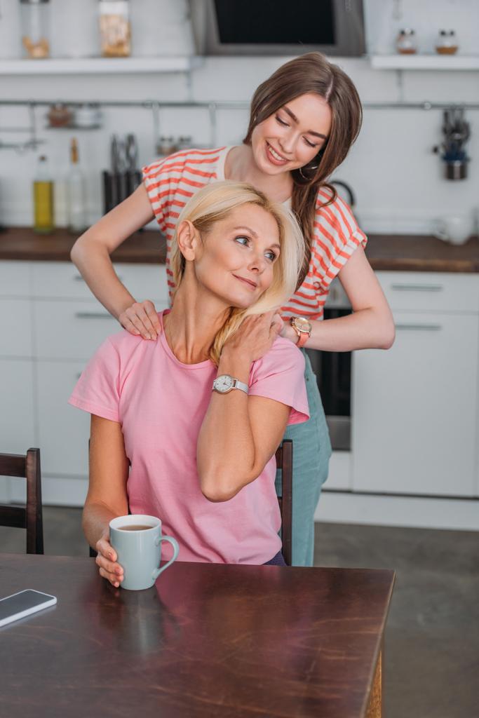 fröhliches Mädchen berührt Schulkinder der Mutter, die mit Teetasse am Küchentisch sitzt - Foto, Bild