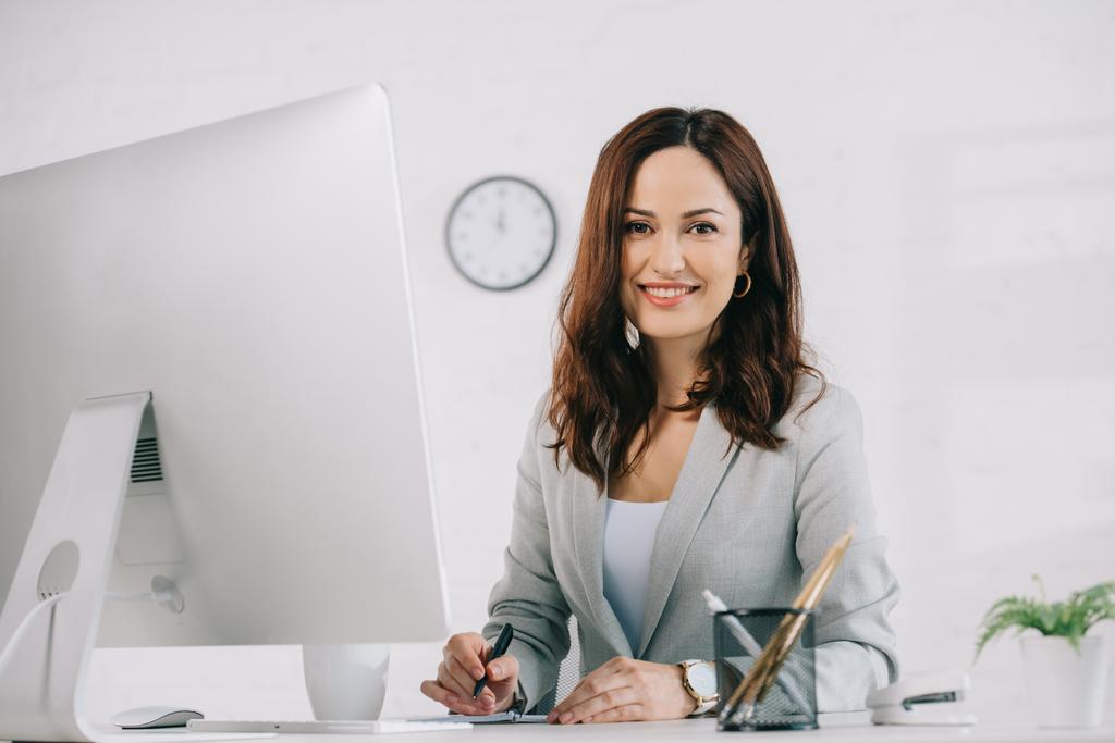 красивая, улыбающаяся секретарша смотрит в камеру, сидя на рабочем месте рядом с монитором компьютера
 - Фото, изображение