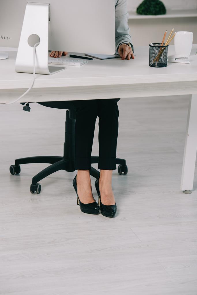 abgeschnittene Ansicht einer Sekretärin in hochhackigen Schuhen, die am Arbeitsplatz im Bürostuhl sitzt - Foto, Bild