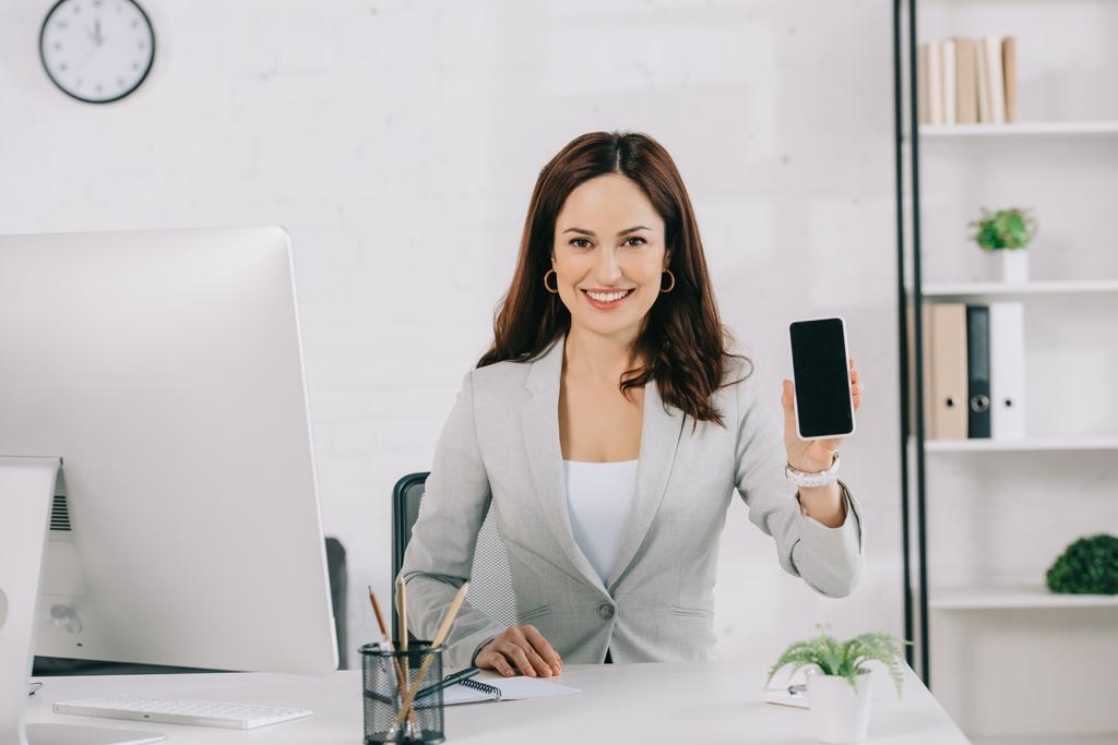 veselá sekretářka ukazující smartphone s prázdnou obrazovkou při sezení na pracovišti - Fotografie, Obrázek