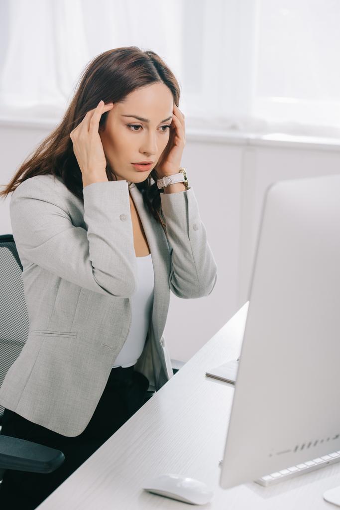 uitgeput secretaresse houden handen in de buurt van het hoofd tijdens het zitten op het werk en lijden aan hoofdpijn - Foto, afbeelding