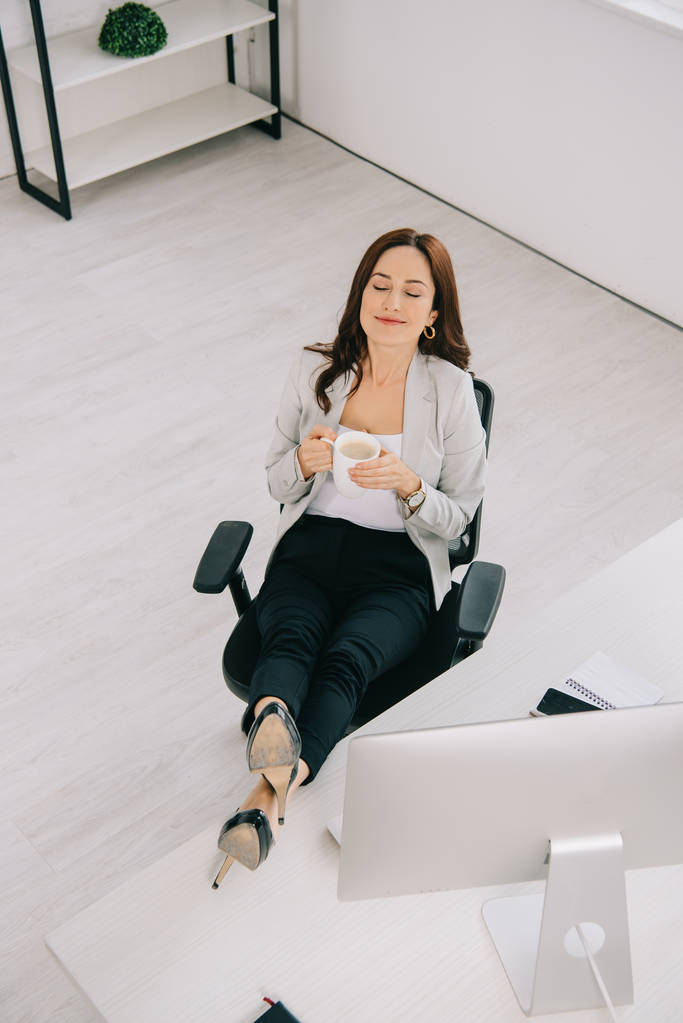 vista de ángulo alto de la secretaria feliz y elegante sentada cerca del lugar de trabajo con los ojos cerrados y sosteniendo la taza de café
 - Foto, Imagen