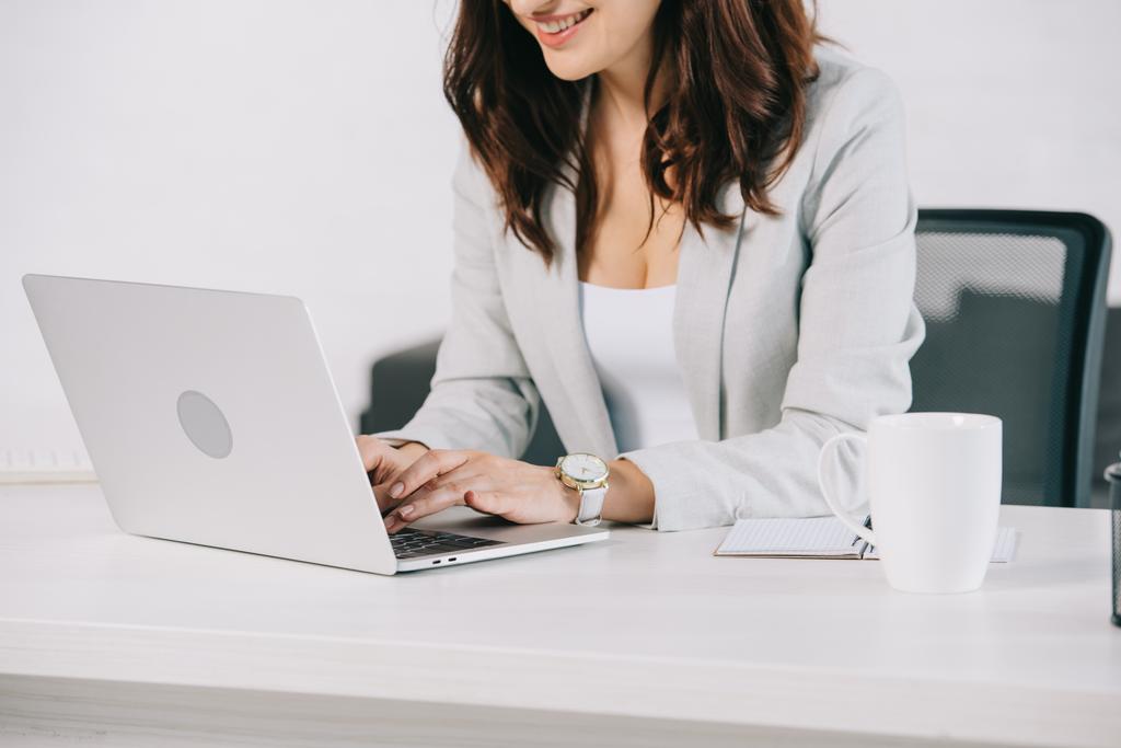 vista parcial de la secretaria sonriente usando el ordenador portátil mientras está sentado en el lugar de trabajo en la oficina
 - Foto, imagen