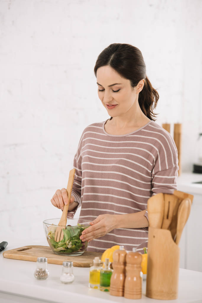 красивая молодая женщина смешивает свежий овощной салат, стоя за кухонным столом
 - Фото, изображение