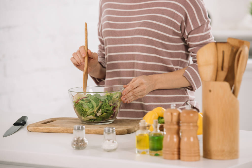 キッチンテーブルに立ちながら新鮮な野菜のサラダを混ぜる女性のクロップドビュー - 写真・画像