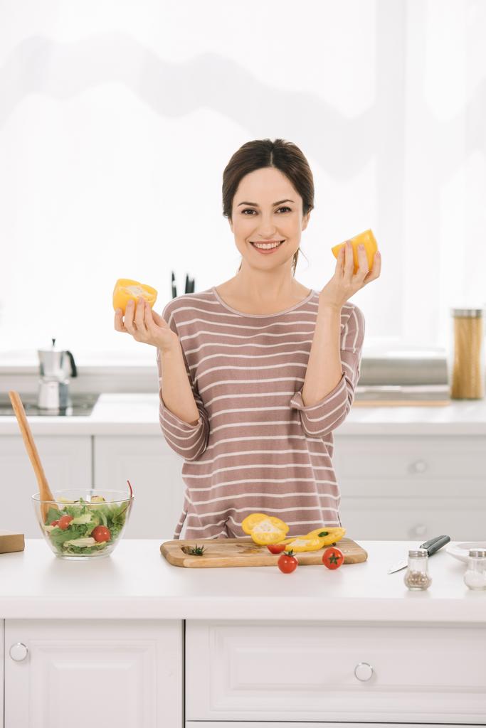Neşeli genç bir kadın mutfak masasının yanında elinde taze sebzeler ve kesilmiş dolmalık biber tutarken kameraya bakıyor.  - Fotoğraf, Görsel