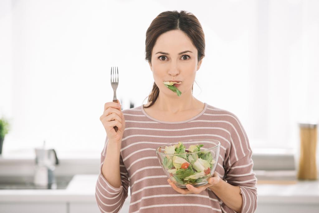 attraktive junge Frau blickt in die Kamera, während sie Gabel hält und frischen Gemüsesalat isst  - Foto, Bild