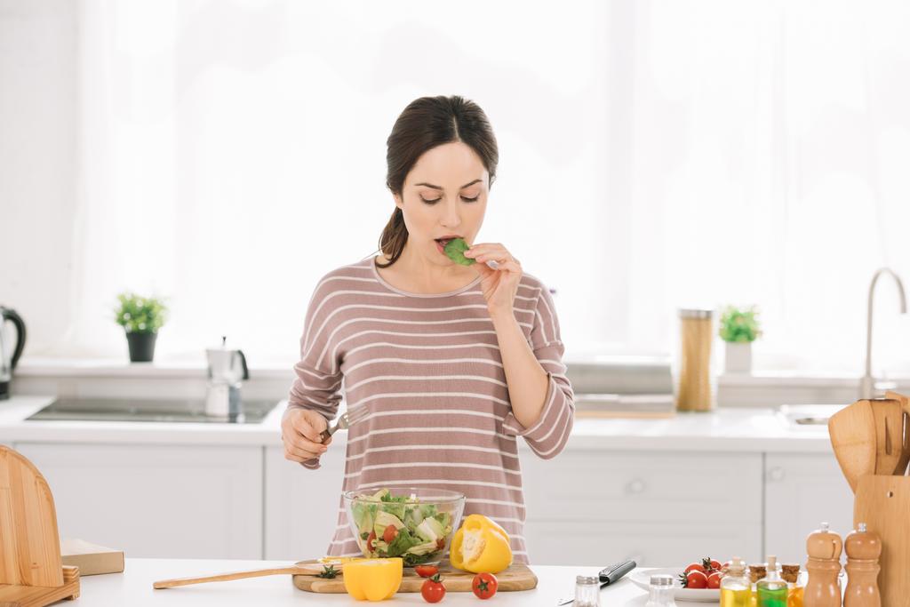 bonita, joven mujer degustación de ensalada, mientras que de pie en la mesa de la cocina con verduras frescas
 - Foto, imagen
