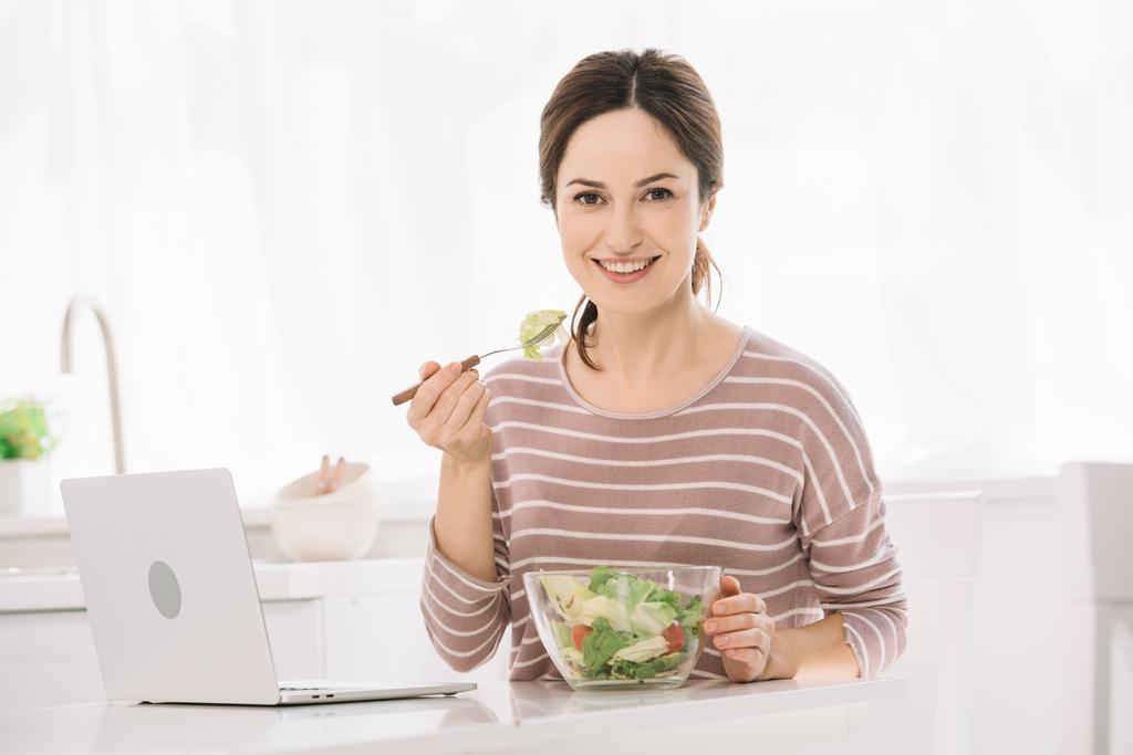 jeune femme heureuse regardant la caméra alors qu'elle était assise à table près d'un ordinateur portable et mangeant de la salade de légumes
 - Photo, image
