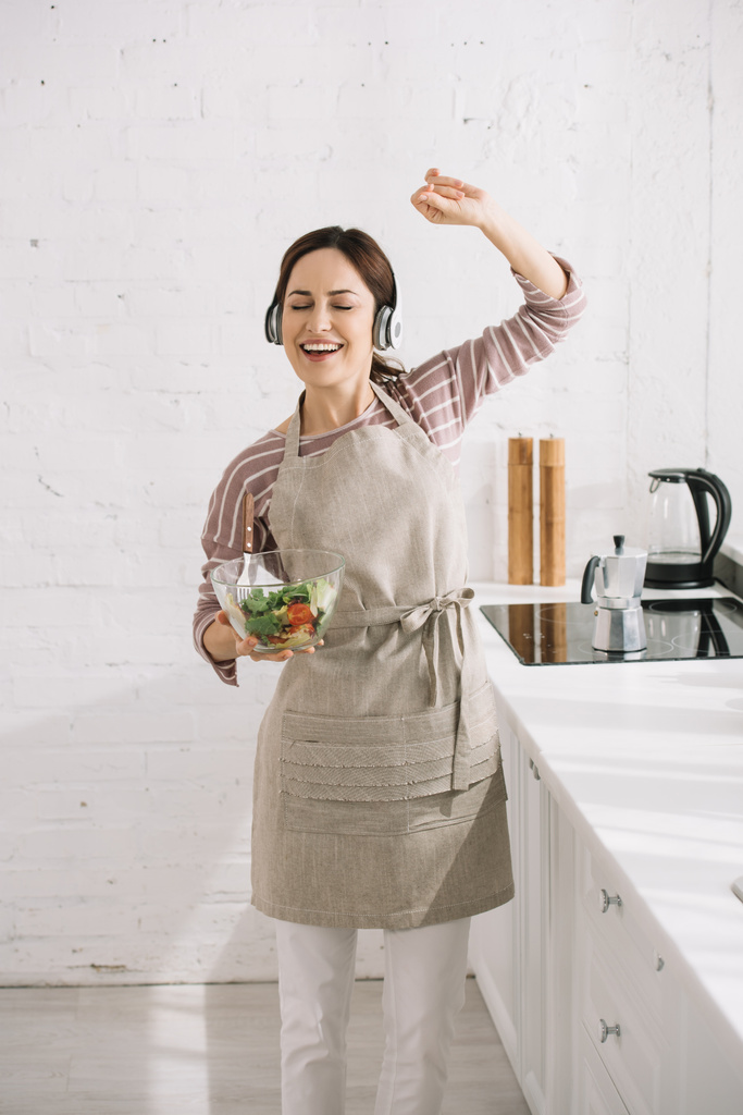 mujer joven feliz en auriculares bailando y cantando mientras sostiene el tazón con ensalada
 - Foto, imagen