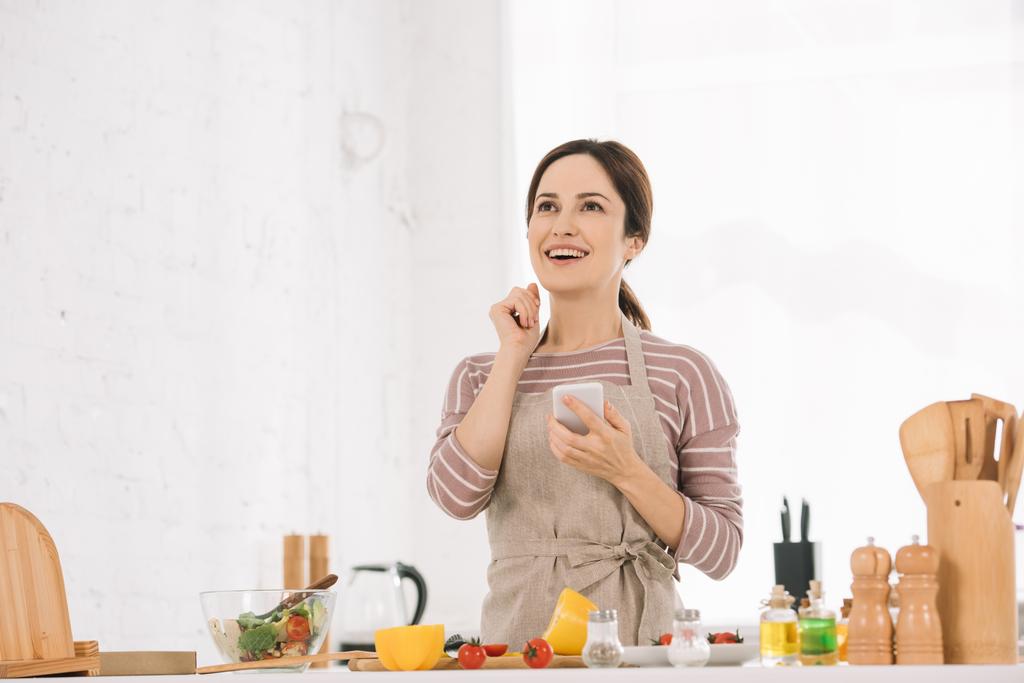feliz joven en delantal sosteniendo teléfono inteligente mientras está de pie en la mesa de la cocina cerca de verduras frescas
 - Foto, imagen