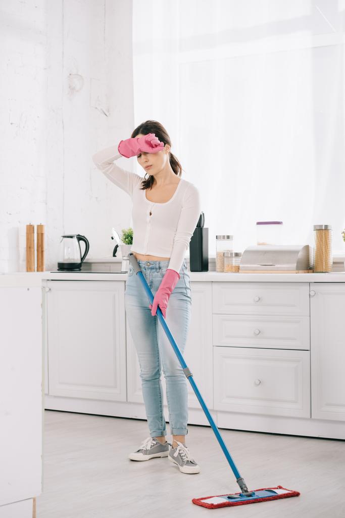 истощенная домохозяйка, стоящая со шваброй на кухне и держащая руку возле головы
 - Фото, изображение