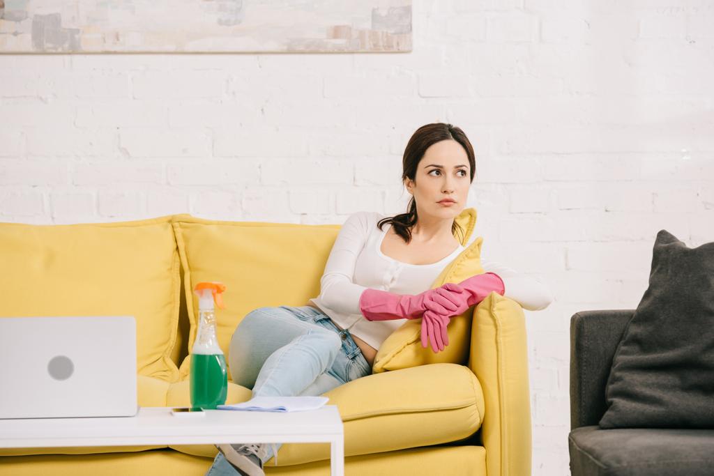κουρασμένη νοικοκυρά κοιτάζοντας μακριά, ενώ κάθεται σε κίτρινο καναπέ κοντά στο τραπέζι με φορητό υπολογιστή και μπουκάλι σπρέι - Φωτογραφία, εικόνα