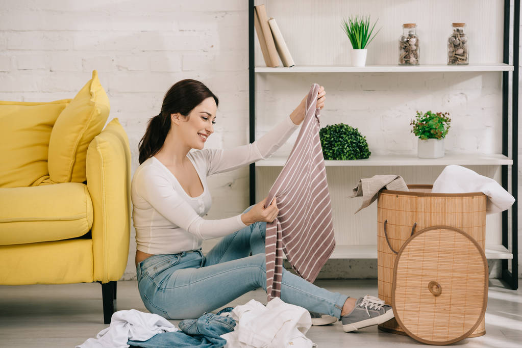 cheerful housewife sitting on floor near laundry basket and holding stripped pullover - Photo, Image