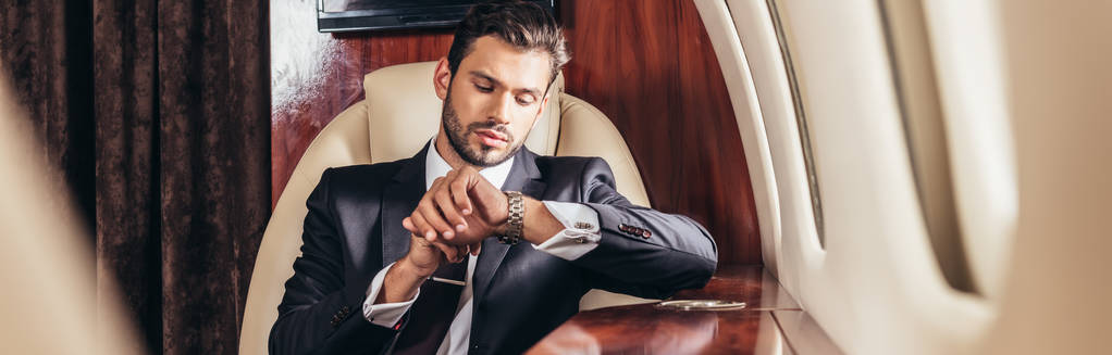 panoramiczne ujęcie przystojnego biznesmena w garniturze patrzącego na zegarek w prywatnym samolocie  - Zdjęcie, obraz