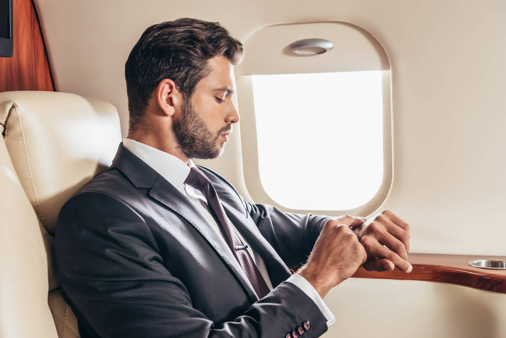 πλαϊνή άποψη του όμορφος επιχειρηματίας με κοστούμι κοιτάζοντας ρολόι χειρός σε ιδιωτικό αεροπλάνο  - Φωτογραφία, εικόνα