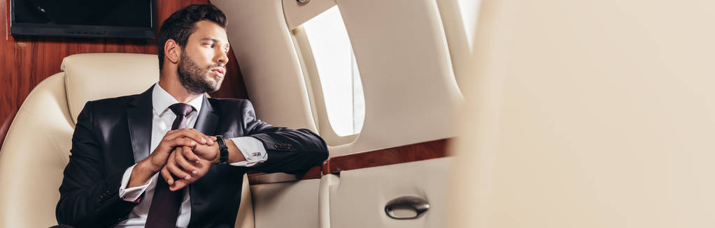 πανοραμική φωτογραφία του όμορφου επιχειρηματία με κοστούμι κοιτάζοντας μέσα από το παράθυρο σε ιδιωτικό αεροπλάνο  - Φωτογραφία, εικόνα