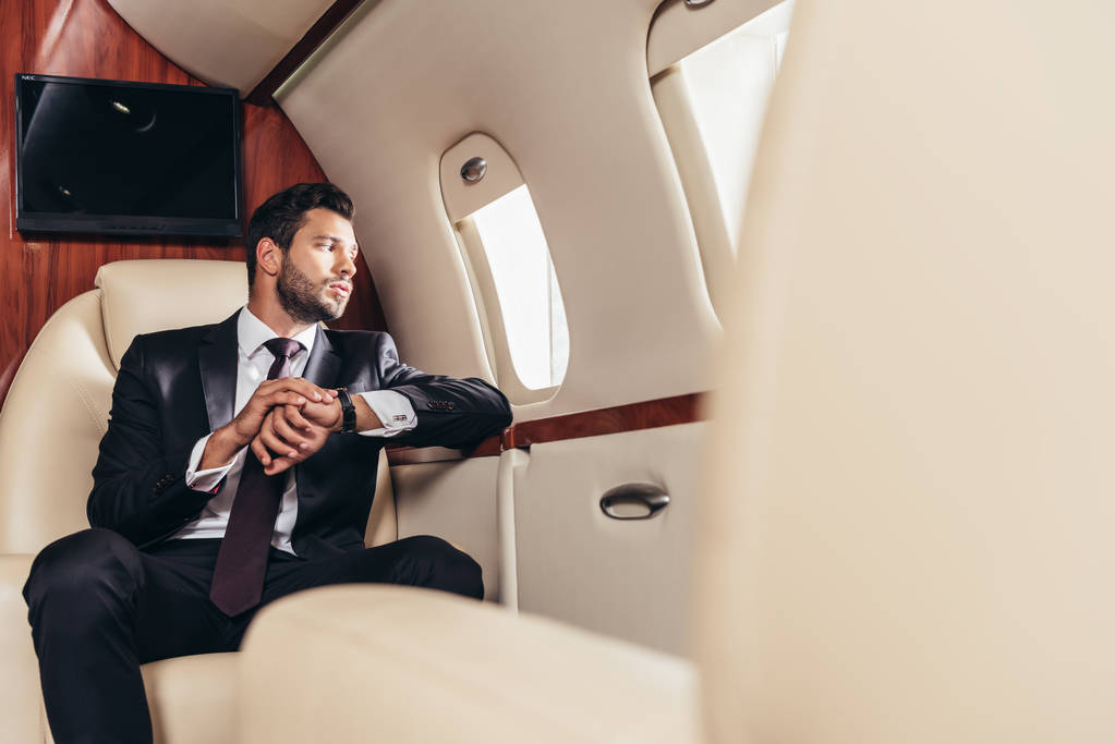 selective focus of handsome businessman in suit looking through window in private plane  - 写真・画像