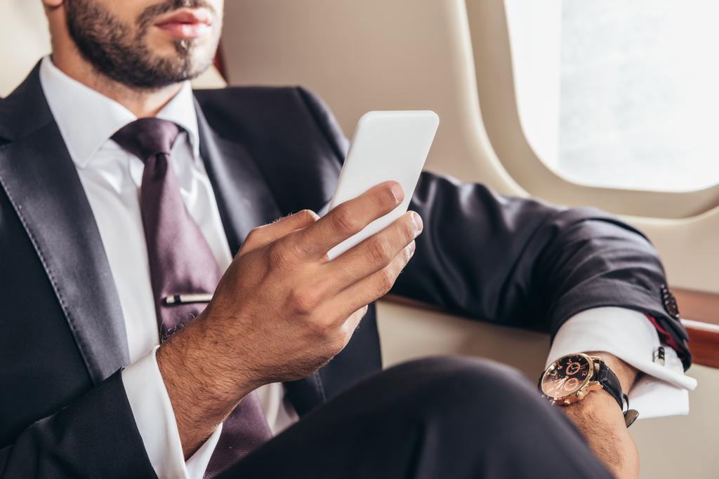 bijgesneden weergave van zakenman in pak met behulp van smartphone in prive-vliegtuig  - Foto, afbeelding