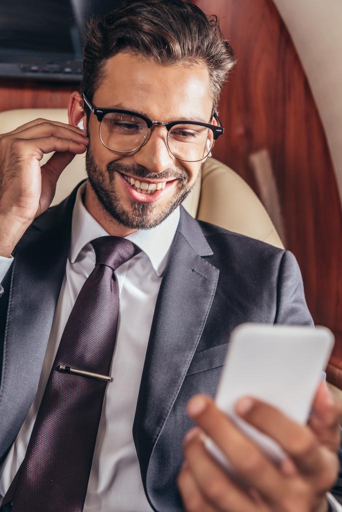 selectieve focus van lachende zakenman die muziek luistert en smartphone gebruikt in privévliegtuigen  - Foto, afbeelding