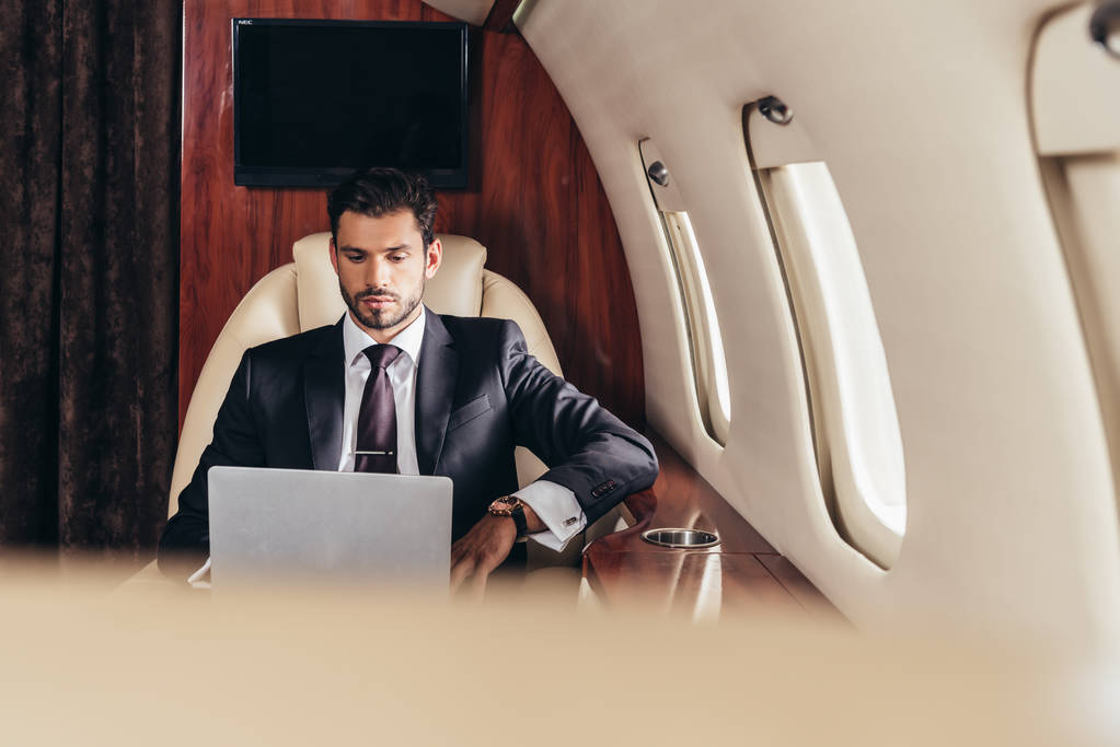 επιλεκτική εστίαση του όμορφου επιχειρηματία με κοστούμι χρησιμοποιώντας φορητό υπολογιστή σε ιδιωτικό αεροπλάνο  - Φωτογραφία, εικόνα