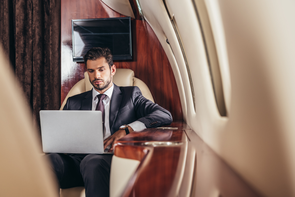 Selektiver Fokus eines gut aussehenden Geschäftsmannes im Anzug mit Laptop im Privatflugzeug  - Foto, Bild