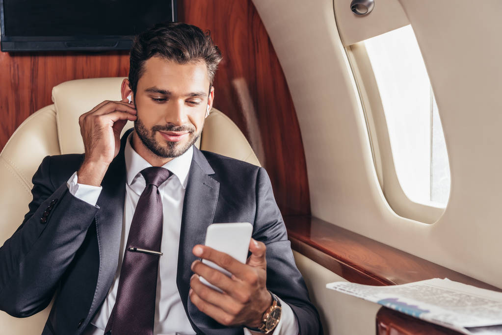 όμορφος επιχειρηματίας με κοστούμι ακούγοντας μουσική και χρησιμοποιώντας smartphone σε ιδιωτικό αεροπλάνο  - Φωτογραφία, εικόνα