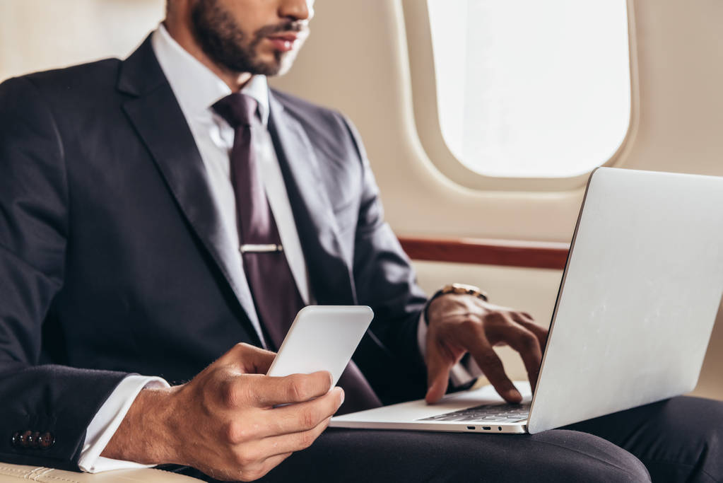 обрезанный вид бизнесмена в костюме с помощью ноутбука и смартфона в частном самолете
  - Фото, изображение