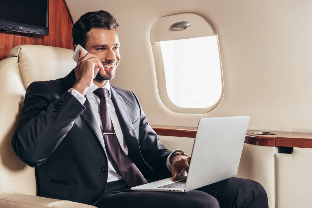 smiling businessman in suit with laptop talking on smartphone in private plane  - Photo, Image