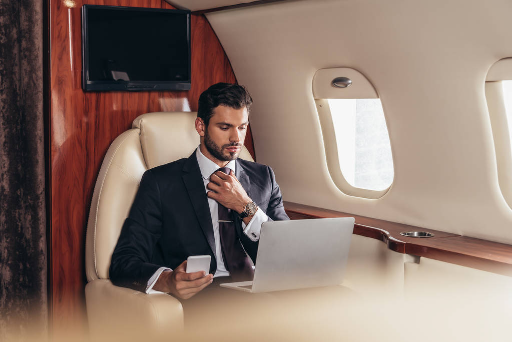 selective focus of businessman in suit using laptop and holding smartphone in private plane  - 写真・画像