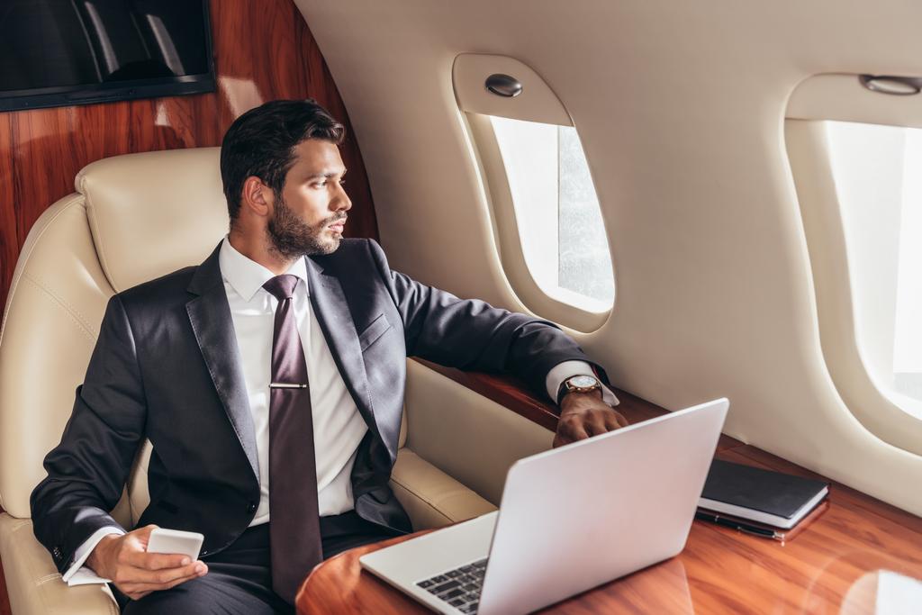 schöner Geschäftsmann im Anzug, der durch das Fenster schaut und sein Smartphone im Privatflugzeug hält  - Foto, Bild