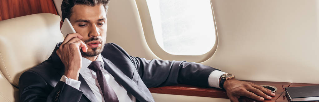 panoramiczne ujęcie przystojnego biznesmena w garniturze rozmawiającego na smartfonie w prywatnym samolocie  - Zdjęcie, obraz