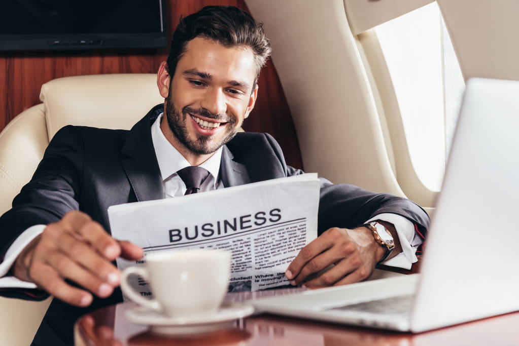 επιλεκτική εστίαση του χαμογελαστού επιχειρηματία στο κοστούμι με εφημερίδα λαμβάνοντας κύπελλο στο ιδιωτικό αεροπλάνο  - Φωτογραφία, εικόνα
