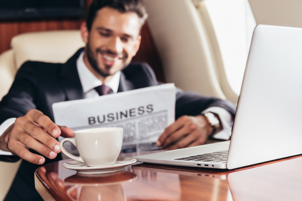 selective focus of smiling businessman in suit with newspaper taking cup in private plane  - Photo, Image