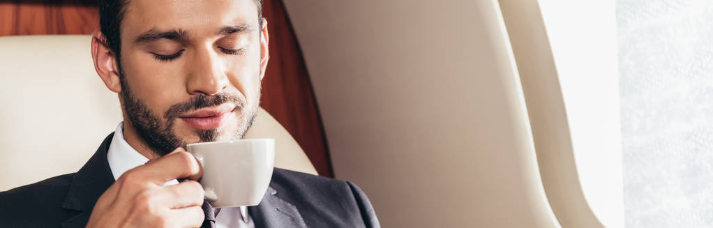 πανοραμική φωτογραφία του όμορφου επιχειρηματία με κοστούμι μυρίζοντας φλιτζάνι καφέ σε ιδιωτικό αεροπλάνο  - Φωτογραφία, εικόνα