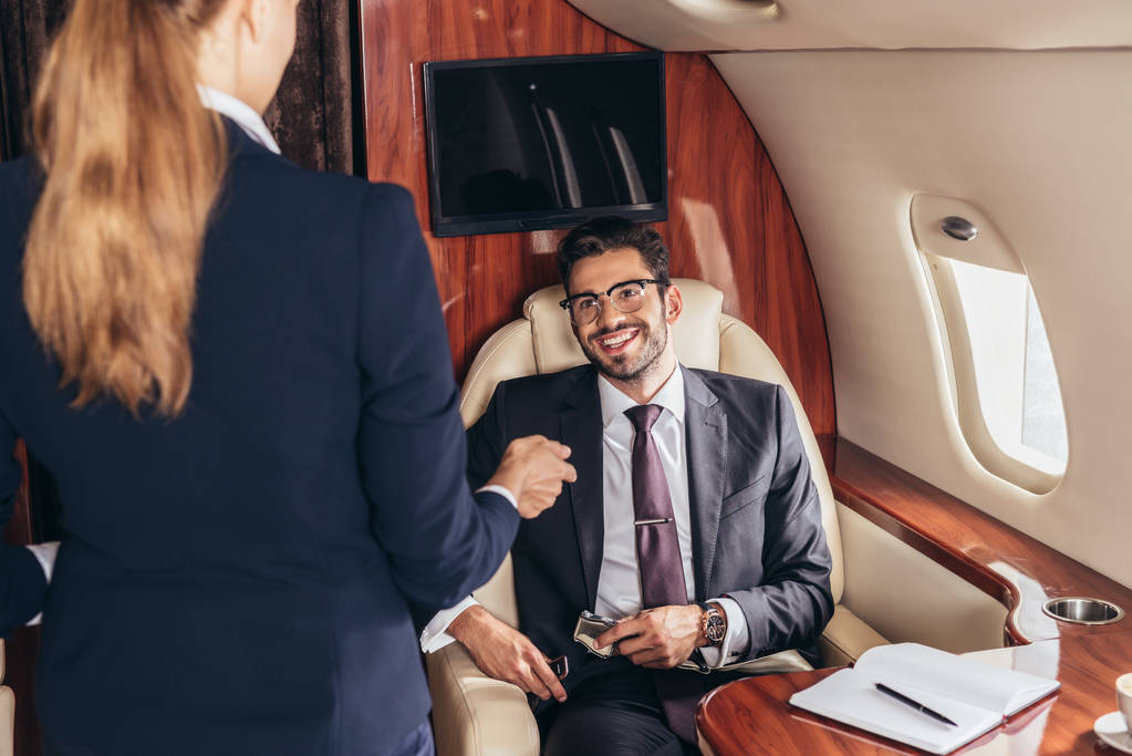 Lächelnder Geschäftsmann im Anzug schaut Flugbegleiterin im Privatflugzeug an  - Foto, Bild