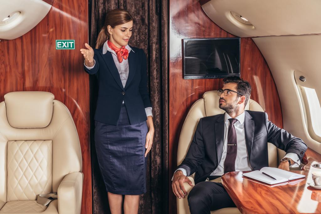 Flugbegleiterin zeigt hübschen Geschäftsmann im Anzug im Privatflugzeug Gesten  - Foto, Bild
