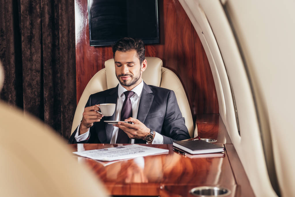 selectieve focus van knappe zakenman in pak die koffie drinkt in privévliegtuig  - Foto, afbeelding